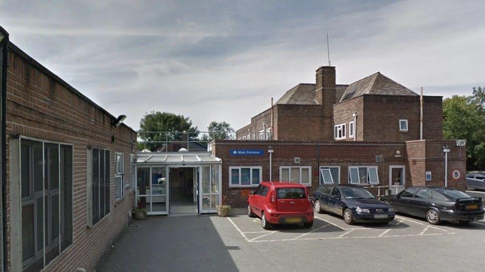 The outside of a brick hospital, with cars parked and a blue Main Entrance sign.