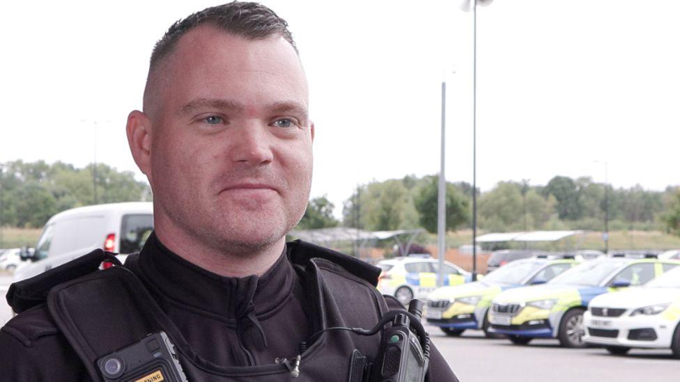 PC Ashley Aspinall in uniform smiling with police cars in the background