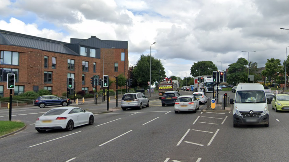 Neville's Cross in Durham is busy with vehicles at the junction on the A167. The traffic lights are on green and vehicles are heading through them. A three-storey brick building - which could be flats or offices such is the relative blandness of the design - with a zinc or copper-style pointed roof is to the left of the junction.