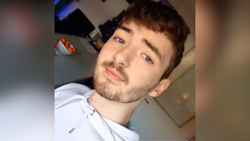 Lee Gannon is a young man with brown hair and some facial hair. He's wearing a light coloured hoodie and in a kitchen. There is a bowl of oranges on the counter.