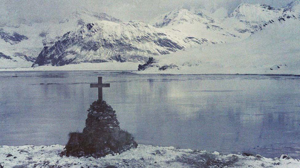 A large cross on top of a stone cairn on the water's edge, with water and snow-covered mountains in the background