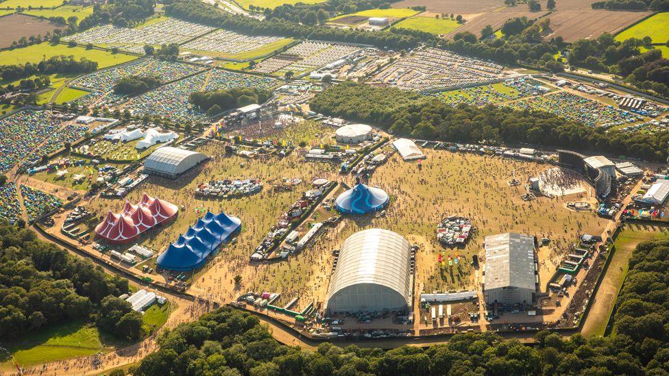 An aerial view of the whole Creamfields site, showing the blue and red big tops and open-air stages surrounded by a fence and a number of campsites filled with multi-coloured tents and areas of dense woodland and farm fields