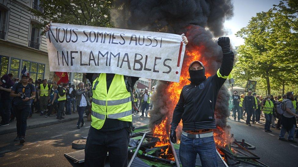 Gilets jaunes (yellow-vests) are seen standing in front of a burning barricade with a banner reading 'we can also burn' on 20 April