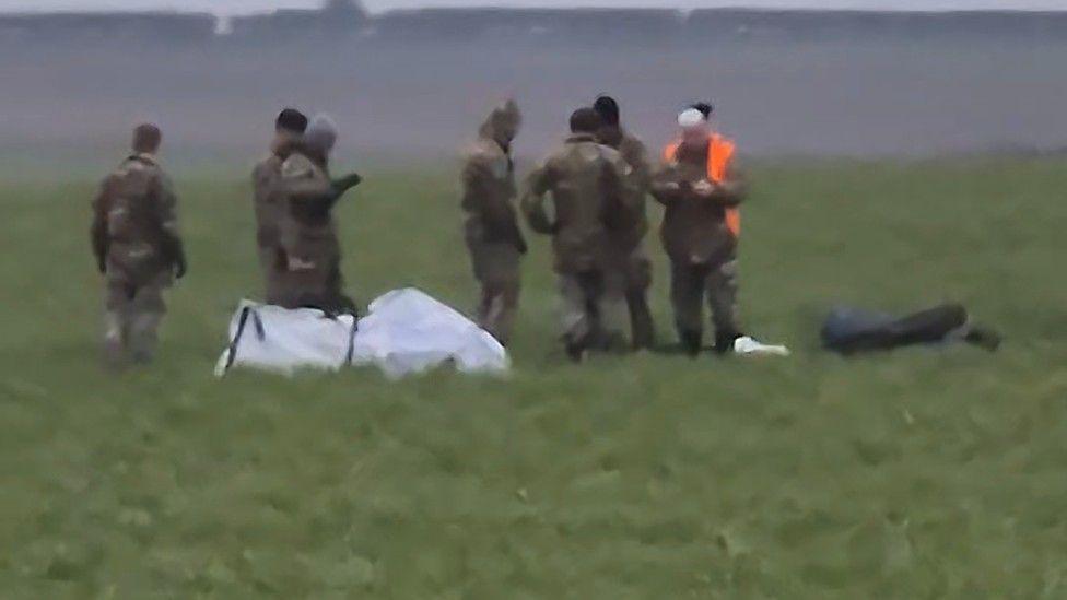 Uniformed crews retrieving the parts. They have been covered in a white tarpaulin. One of the team is wearing an orange hi-vis jacket.