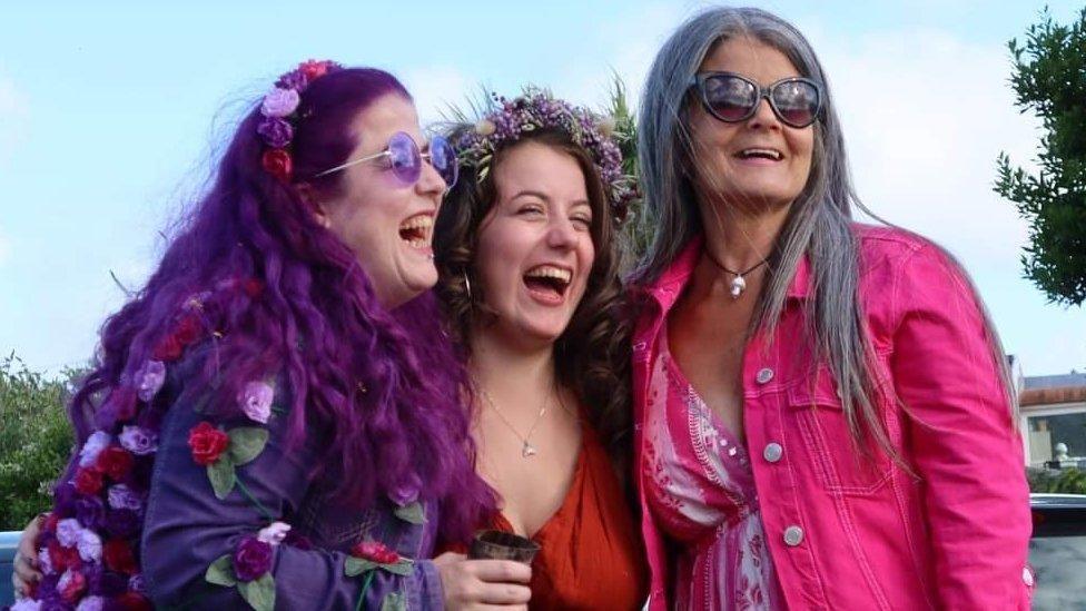 Claire Lewis and her two adult daughters wearing bright pink, red and purple dresses and laughing with their arms round each other