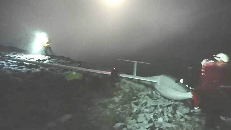 A glider which has crashed atop of the Cheviot. The weather conditions are poor and there are two rescue members in the shot. 