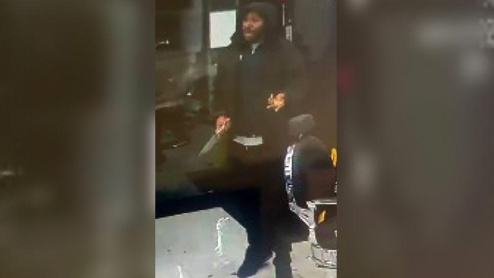 A man stands in a barbers' shop dressed in black with the hood of his coat over his head. He is gesticulating with one hand and the other one has a knife in it. He has a blue sweater on.