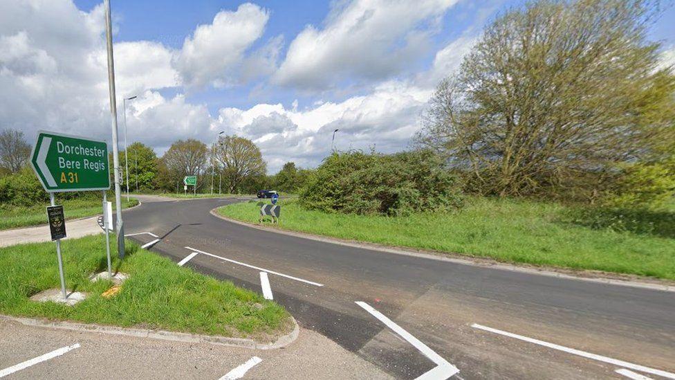 Roads leading on to roundabout with green and white signs trees and shrubs  are growing on the grassed roundabout.