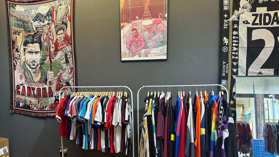 The inside of the couple's vintage football shirt shop in Manchester. There are two racks containing lots of different coloured football shirts in front of a dark grey wall. There is also a mural or Eric Cantona on the wall to the left and in the middle there is one of Lionel Messi which also has the World Cup trophy on it. To the left you can see half of another black and white mural about the former player Zinedine Zidane