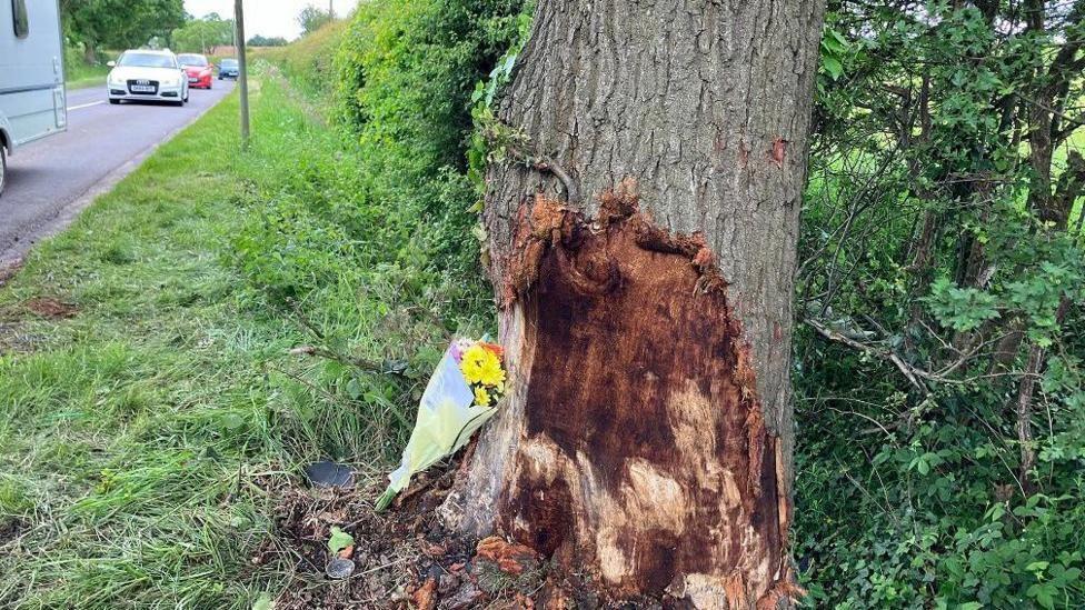 The damaged tree, with grass around it and a boquet of flowers laid beside it