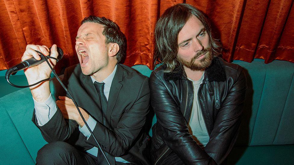 The two members of the band Benefits, Kingsley Hall and Robbie Major, sitting on a teal-coloured sofa in front of a rust-coloured velvet curtain. Kingsley Hall is holding a microphone in front of his face and appears to be screaming or singing loudly - or pretending to. Robbie Major is looking away, pensively. Kingsley Hall is wearing a black suit, grey shirt and black tie. Robbie Major is wearing a black leather jacket over a grey t-shirt. 