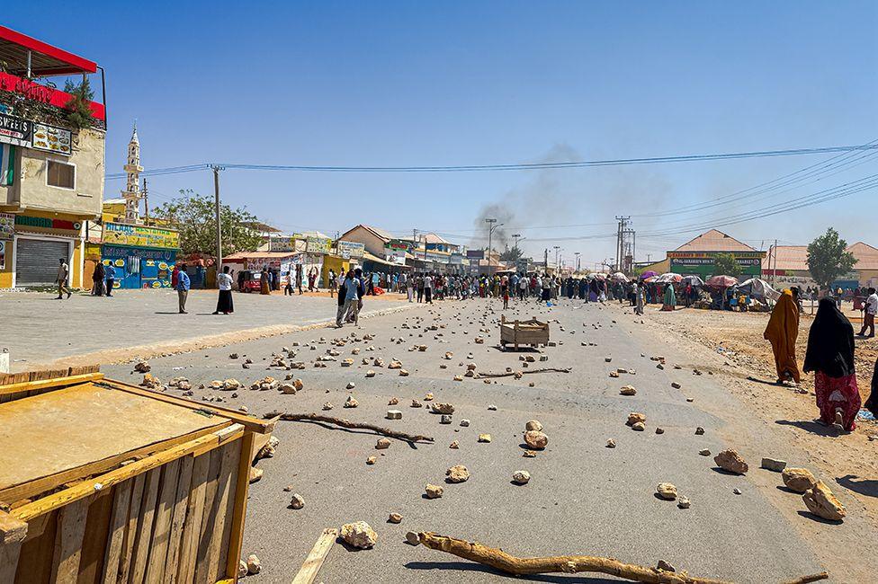 A tarmac road is in the foreground strewn with wooden sticks and stones as a result of a protest. A crowd can be seen in the distance, where a small cloud of smoke is rising.