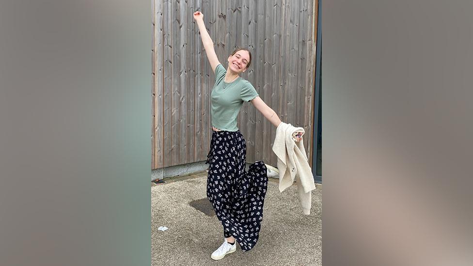 Eleanor stands in a courtyard setting in front of a timber-panelled wall. She's doing a celebration pose, one arm stretched out high above her head and the other pointing towards the ground. One white-trainered foot is off the ground, cocked up behind her. She's wearing a green t-shirt and long, black skirt with a white symbol pattern. She's got her head slightly tilted and is smiling widely.