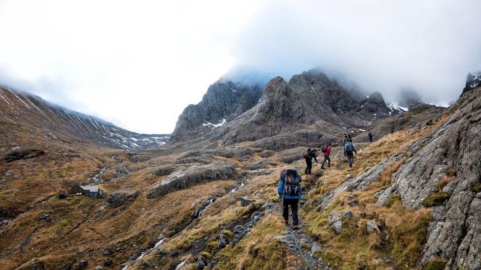 People walking up Ben Nevis. There is lots of rock and grass and people wearing rucksacks.