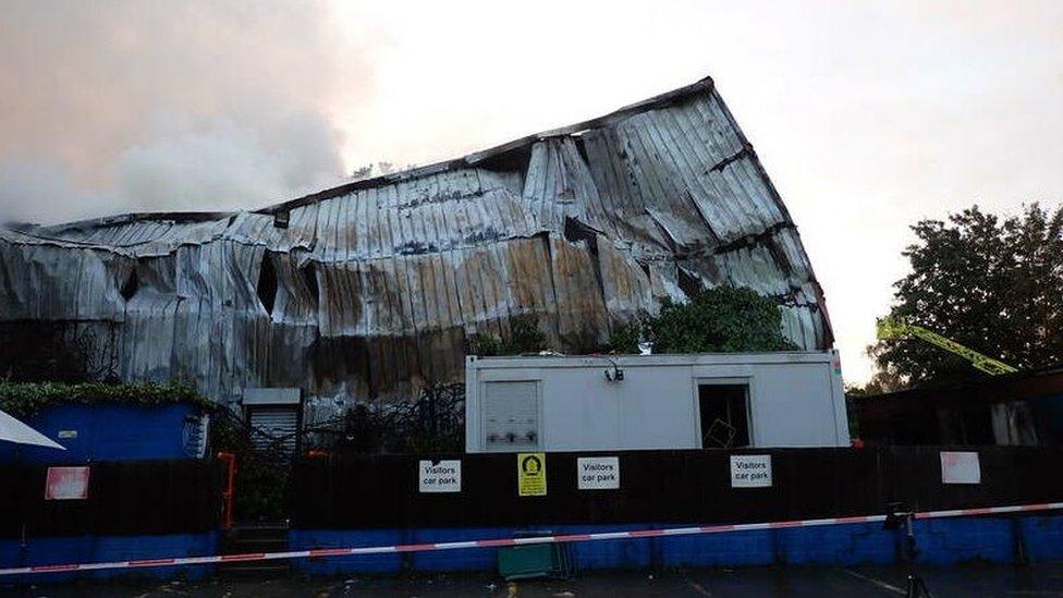 Daytime picture showing a partially-collapsed metal warehouse with smoke rising from it.