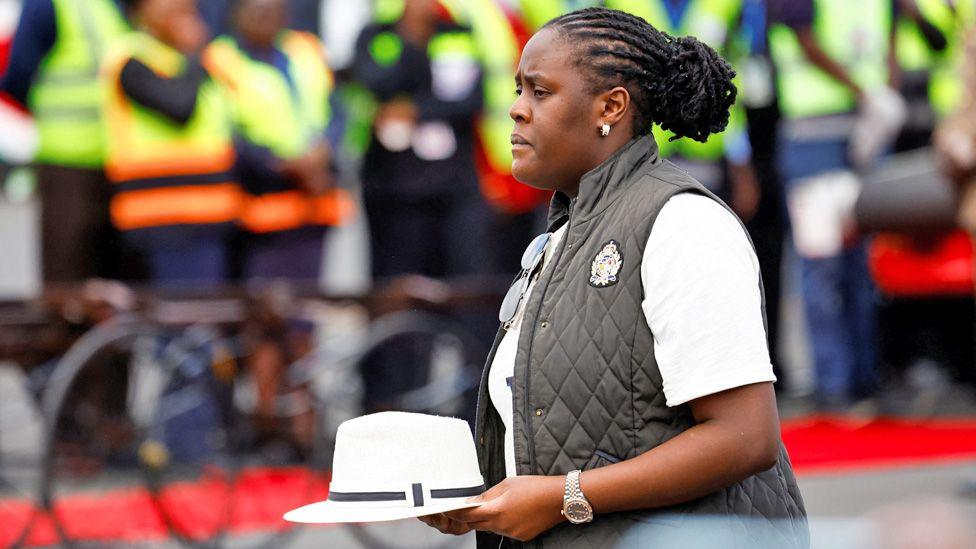 Raila Odinga's daughter Winnie Odinga, in a white T-shirt and black waitcoat, carries his hat as she arrives at Jomo Kenyatta International Airport in Nairobi. Behind her officials dressed in high vis uniforms can be seen.
