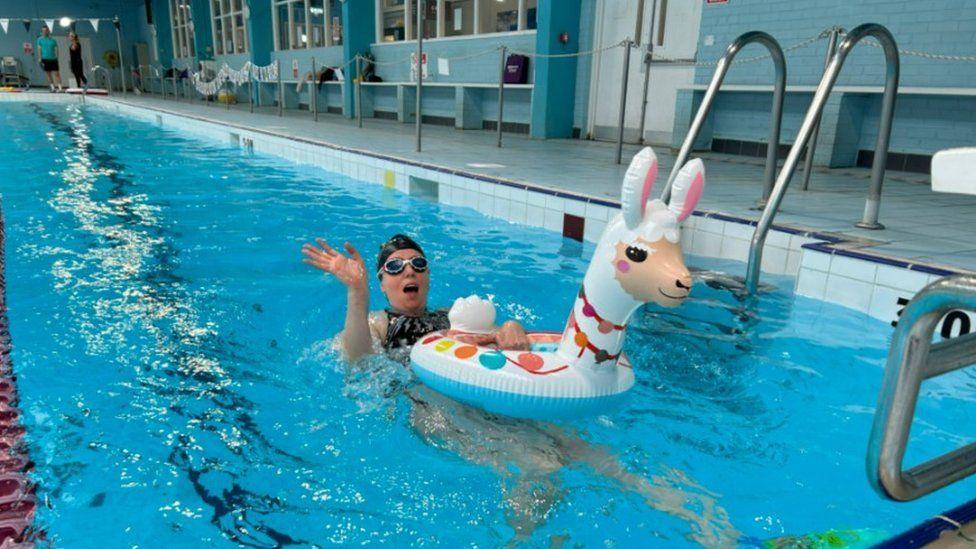 Woman waving from pool wearing a black swim hat and goggles she is swimming on her back pulling and inflatable Llama.