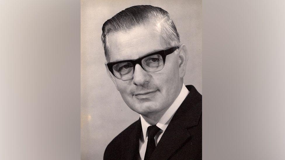 Fred Martin pictured as an older man, wearing a pair of 1960s style glasses with thick frames on top and no frame below. His hair is slicked to one side and he is looking just off camera with a gentle smile. It appears to be in black and white. He is wearing a dark suit and tie and white shirt.