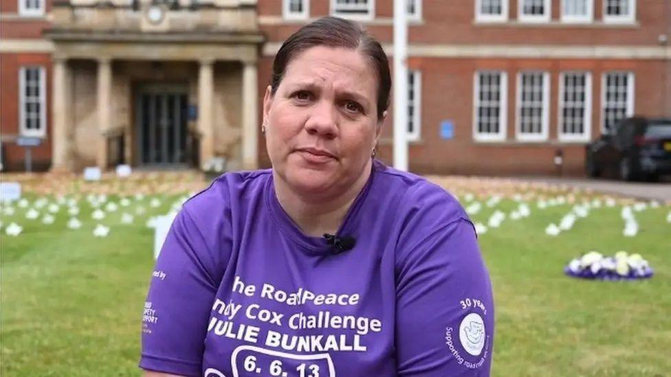 Emilie Bunkall with dark hair tied back. She is looking at the camera and wearing a purple T-shirt with white writing that says "The RoadPeace Andy Cox Challenge Julie Bunkall", together with the date of her mother's death, 6 June 2013.