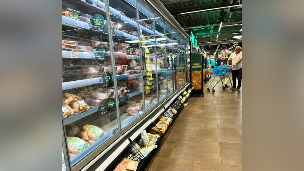 An M&S shopping aisle is lined with fridges, which are behind glass doors. Below the fridges are wired racks containing snacks which stick out.