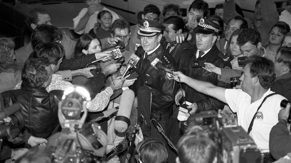 A black and white picture of a police officer giving a press conference with dozens of broadcast journalists surrounding him