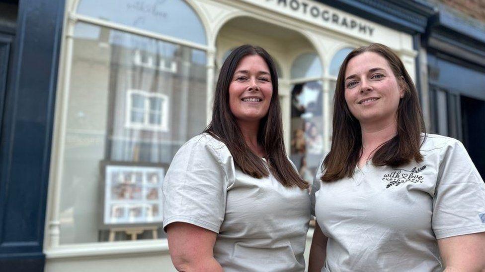 Samantha Cartwright (left) and Victoria Scott standing outside their With Love Photography studio. They both have long dark hair and are looking into the camera. A number of photographic images, including baby portraits, are displayed in the shop windows behind them.