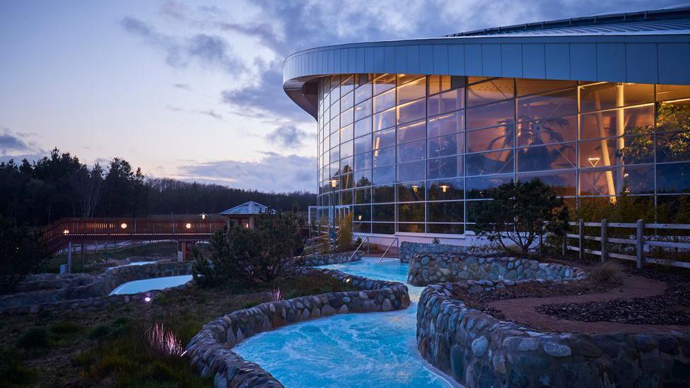 An indoor and outdoor water complex at dusk surrounded by trees