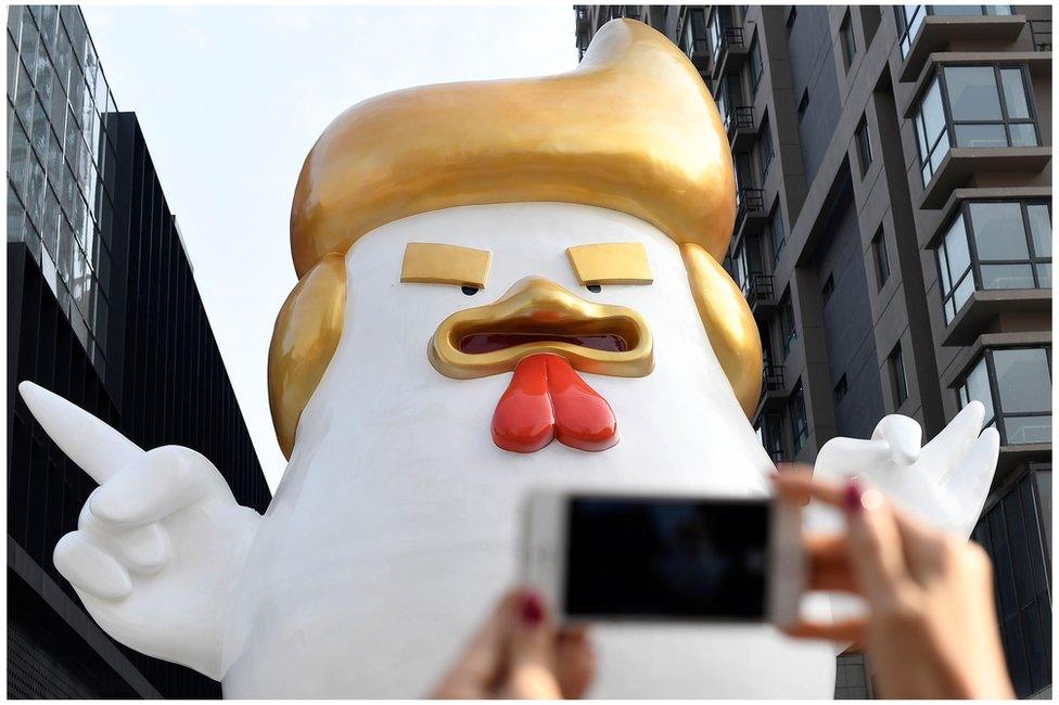 A giant rooster sculpture resembling Donald Trump outside a shopping mall in Taiyuan, Shanxi province, with a mobile phone being held by a woman's hands in the foreground. 24 December 2016.