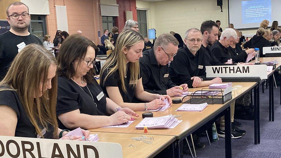 People sitting at rows of tables counting election votes