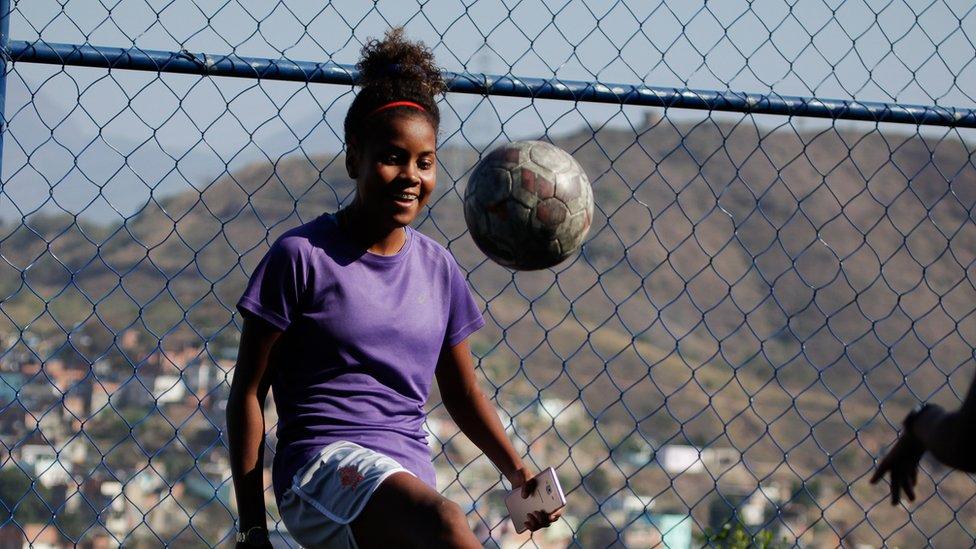 Thaissa controls the ball with a solo kick, in front of a chain-link fence through which the grassy hills beyond the favela can be seen