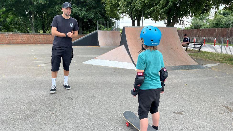 Young boy wearing helmet takes instructions from man in skate park