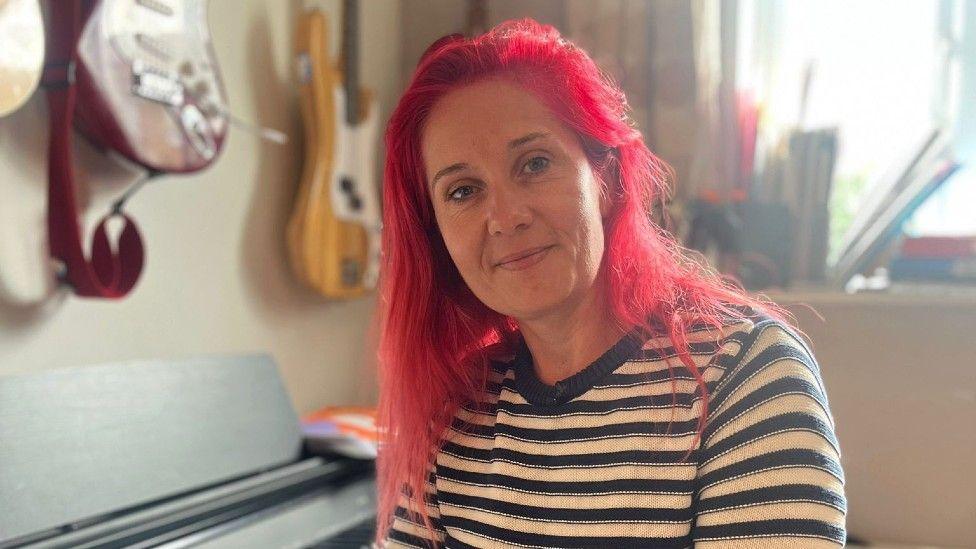 Sharon Stanton is looking into the camera while sat at a piano, with some guitars on the wall behind her. She has long red hair and is wearing a blue and white top.
