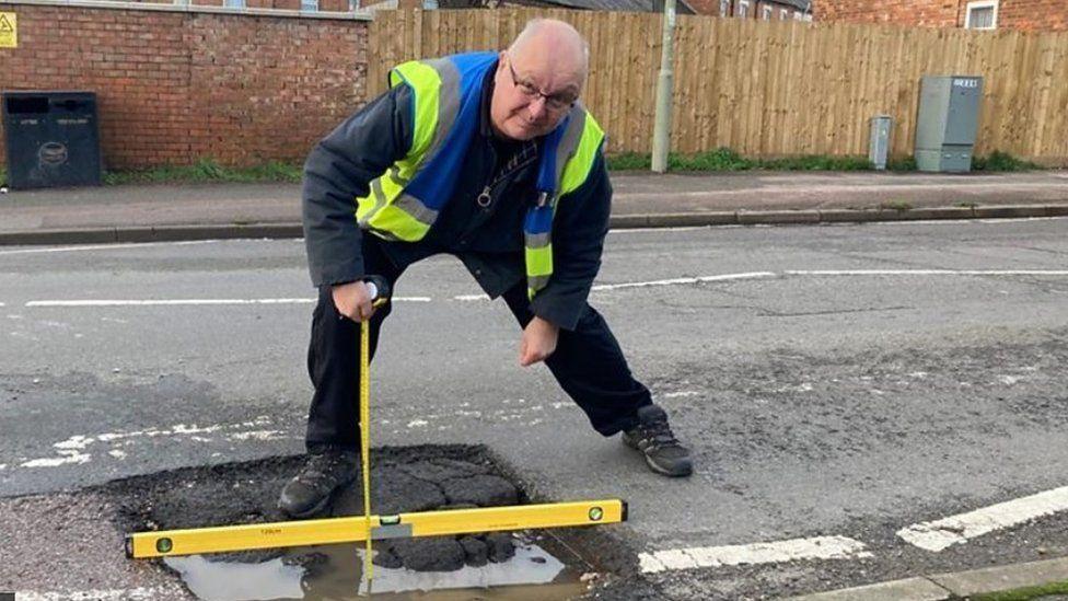 Mark Morrell with short white hair and a yellow and blue hi-vis hacket standing on a road with a yellow measuring device which is checking the width of a large pothole