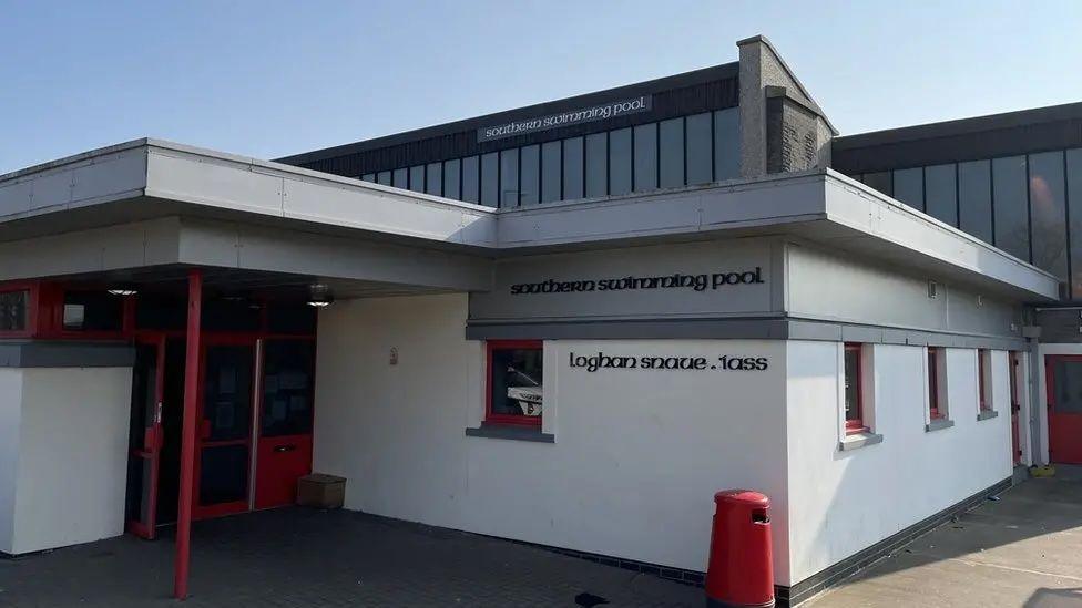 The exterior of the Southern Swimming Pool, a white block-like building, with a sign that reads southern swimming pool, there is a second storey in the background with large windows.