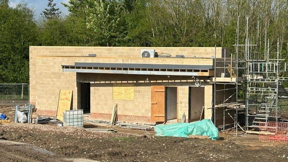 A half-built building in honey-coloured blocks with a flat roof, scaffolding to one end, and bare earth around it. 