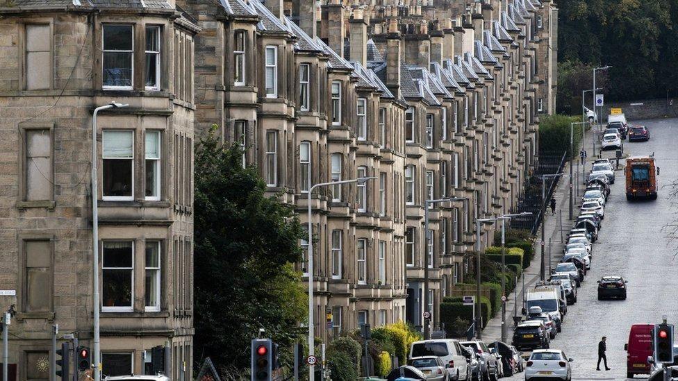 A row of tenement flats on a hill with a line of parked cars outside. A lone man can be seen crossing the road