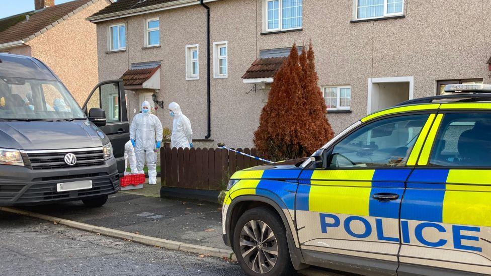 Two people in white forensic suits, pictured outside a brown terraved house. They are stood beside a large, grey van, with police table across a wooden fence. A white, blue and neon yellow police car is also parked outside the properties. 