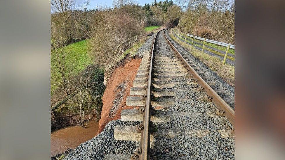 A picture of a railway track, where the embankment on the left-hand-side has fallen away in a landslip. A river can be seen below this.