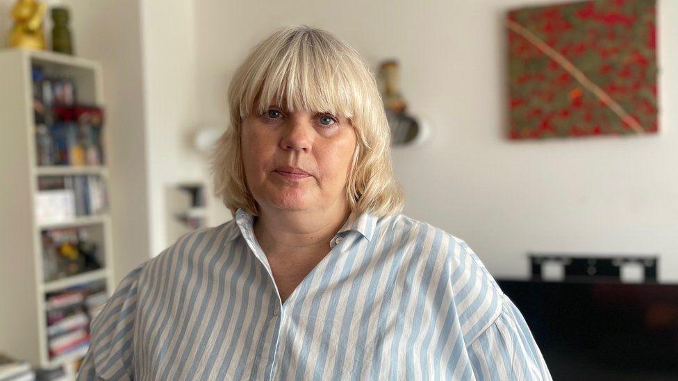 Justine Gordon-Smith stands in a room and has an unsmiling expression as she faces the camera. She has shoulder-length blonde hair with a fringe and wears a shirt with vertical blue and white stripes.