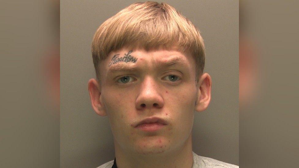 A police photo of a man with short blond hair looking seriously into the camera with a frown. He has a black tattoo that reads "freedom" above his eyebrow.