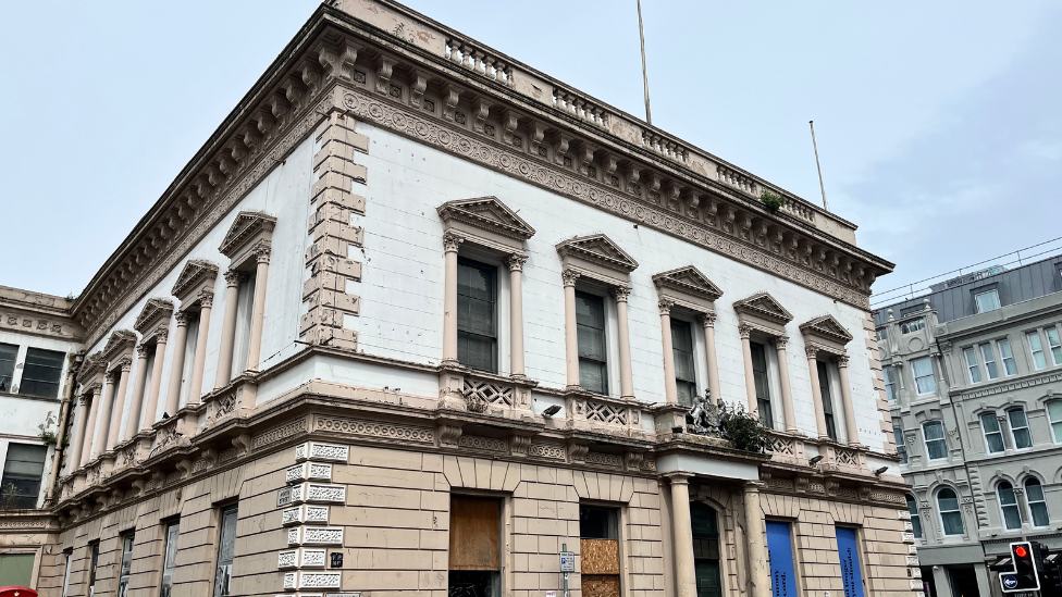 Exterior of large stone, derelict building. It has numerous ornate windows, some of which are boarded up with wood and blue signs