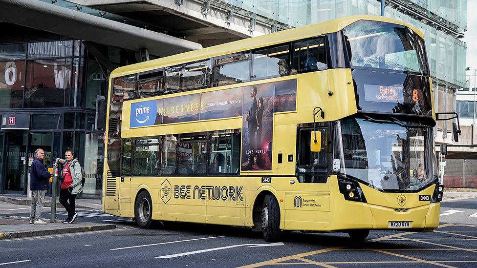 Yellow double decker 'Bee Network' branded bus in Manchester city centre parked next to a bus stop with two people standing next to it, in Manchester in September 2023