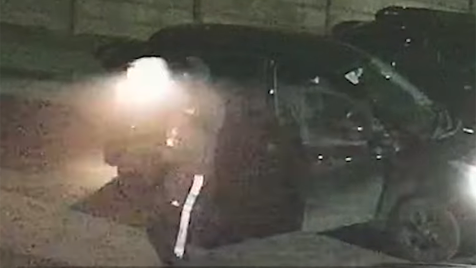 A man standing next to a black car and dressed in dark clothing, with a white stripe down the trousers, is firing a gun. The flash of the shot and smoke can be seen as the weapon is used.