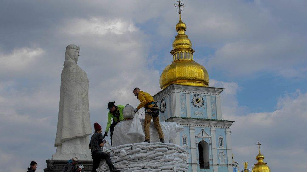 people stand on sandbags piled up high near a statue with a golden topped building in the background