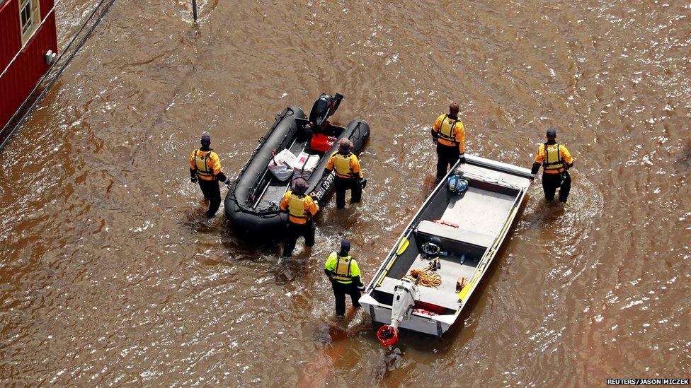 Five men in yellow rescue jackets walking beside rescue boats in floodwaters