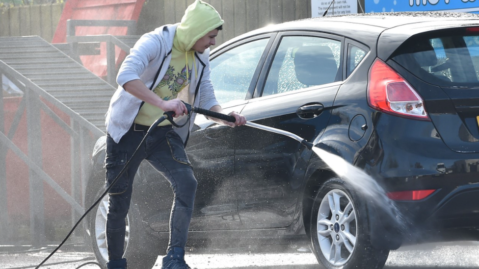 A man in a yellow hooded top using a power hose to clean a car