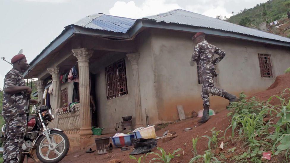 Two police officers, one with a motorbike, outside a house on a hill in Waterloo in Freetown. The house is made of concrete with a corrugated iron roof and some pots and a few maize plants can be seen outside.