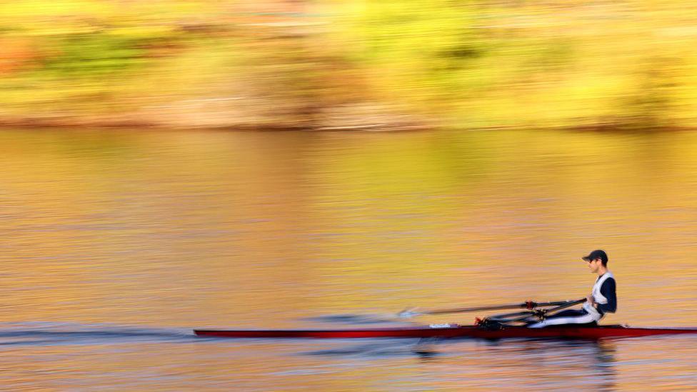 A single sculler rows across calm water in a long, narrow boat, with blurred autumn foliage in shades of yellow and orange reflected on the surface.