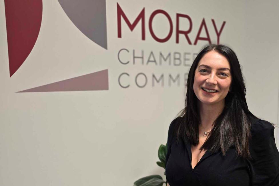 Sarah Medcraf, woman with black hair smiling at camera, wearing black dress, with 'Moray Chamber of Commerce' behind her.