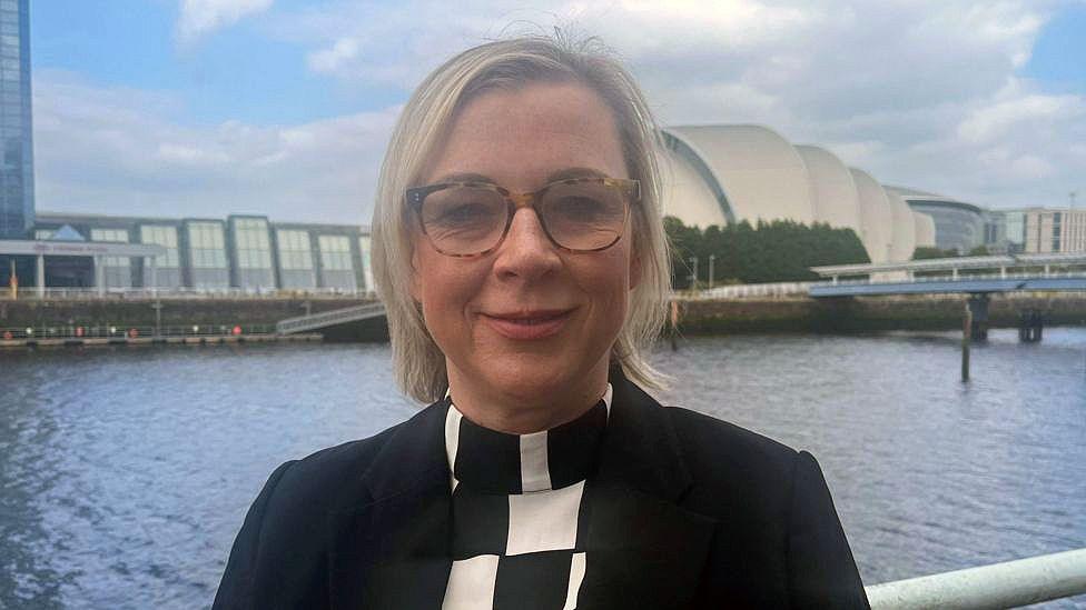 Fiona Dyer, the director of the children and young people’s centre for justice. She has blonde hair and glasses. She is wearing a black jacket and a black and white checked top. Fiona is standing in front of the River Clyde with the Armadillo visible in the background.